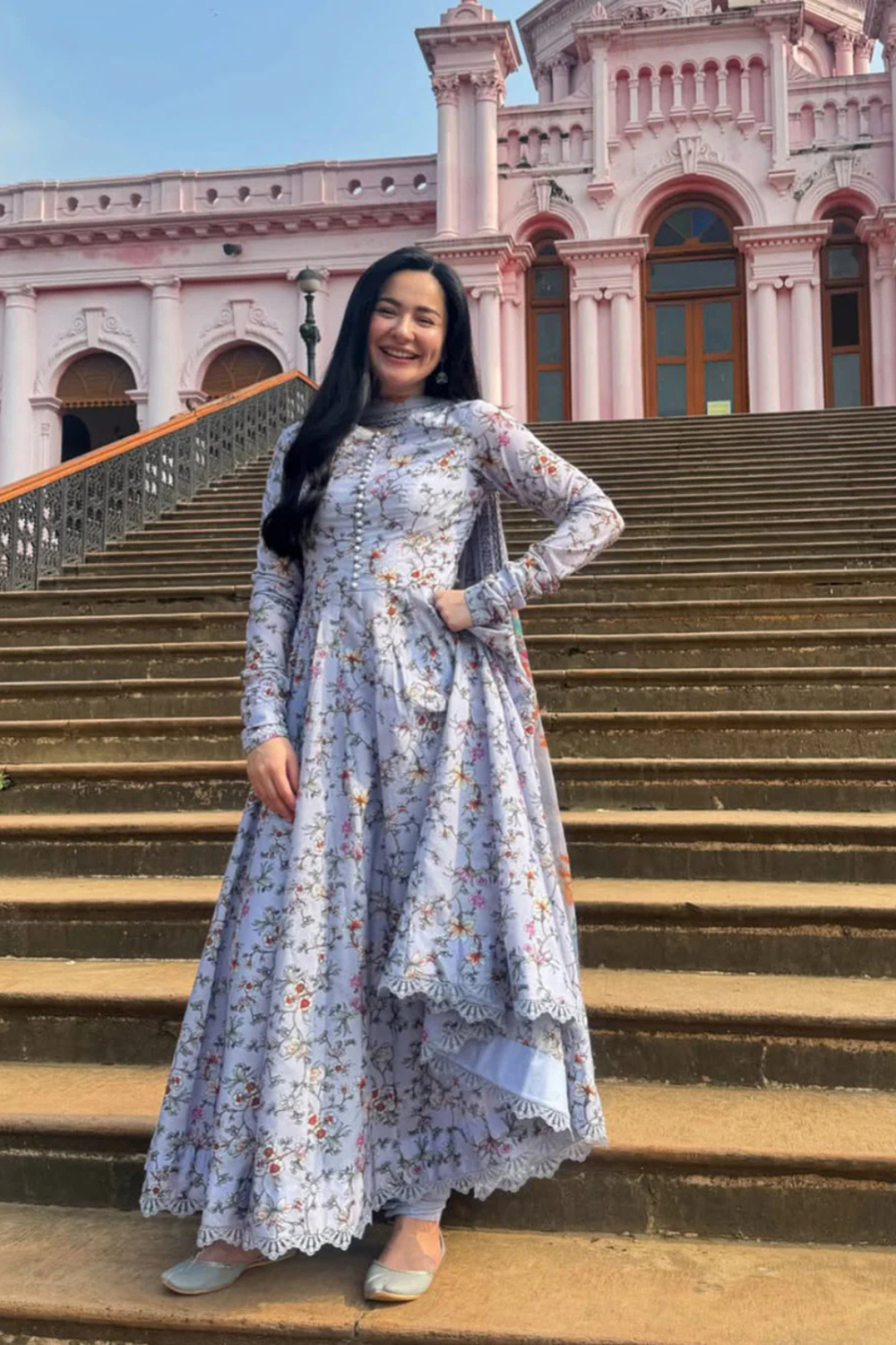 Smiling woman in floral printed light blue traditional dress standing on wide stone steps of a pink historic building