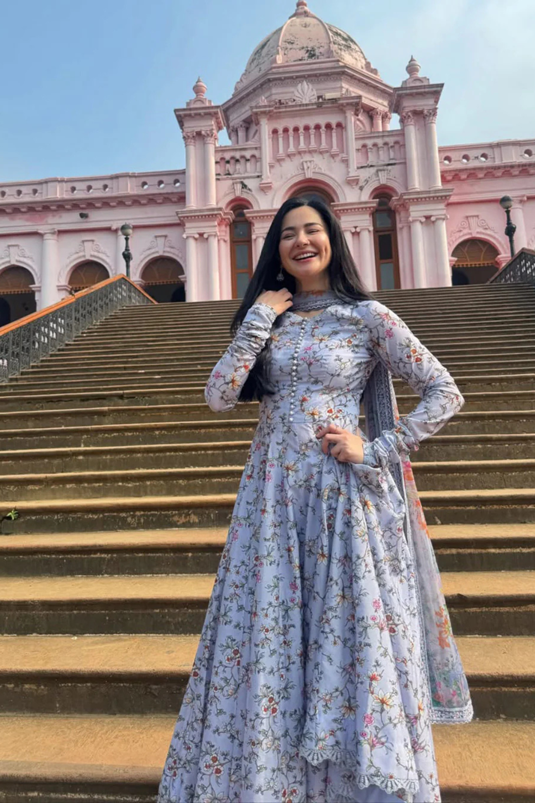 Smiling woman in blue floral anarkali dress standing on stone steps before pink historic building